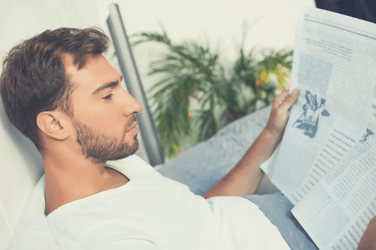 man reading a newspaper in bed with his back resting on a pillow