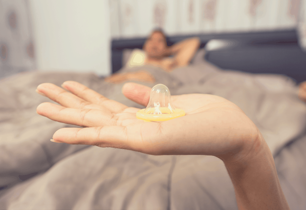 woman holding an unwrapped condom while her partner waits in bed